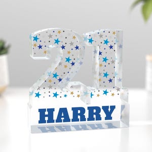 Puede incluir: Un bloque acr&iacute;lico transparente con el n&uacute;mero "21" y el nombre "HARRY" en azul. Los n&uacute;meros est&aacute;n decorados con estrellas azules, doradas y grises. Un objeto decorativo para un cumplea&ntilde;os.