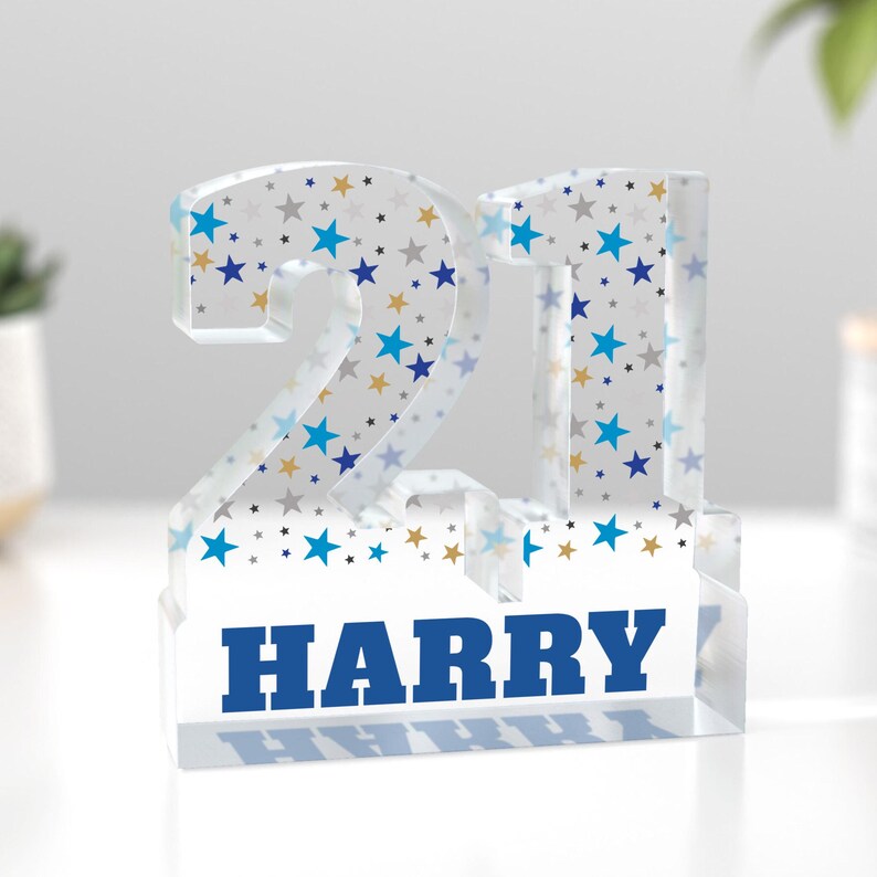Puede incluir: Un bloque acr&iacute;lico transparente con el n&uacute;mero "21" y el nombre "HARRY" en azul. Los n&uacute;meros est&aacute;n decorados con estrellas azules, doradas y grises. Un objeto decorativo para un cumplea&ntilde;os.