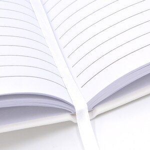 May include: An open notebook with white pages featuring thin black horizontal lines. A white ribbon bookmark is positioned vertically down the center of the open pages, extending from the spine of the white cover of the notebook.