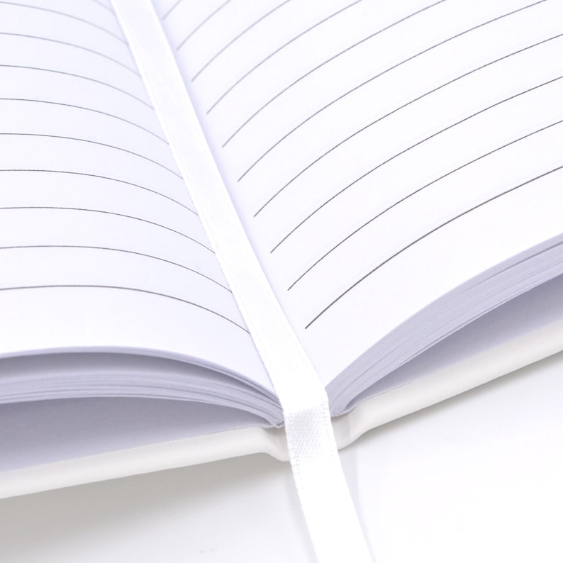 May include: An open notebook with white pages featuring thin black horizontal lines. A white ribbon bookmark is positioned vertically down the center of the open pages, extending from the spine of the white cover of the notebook.