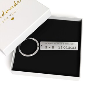May include: A silver rectangular keychain with a keyring, presented in a white gift box with a black interior. The keychain is engraved with the text "It started with a message..." and initials S &hearts; M, along with the date 12.09.2023.
