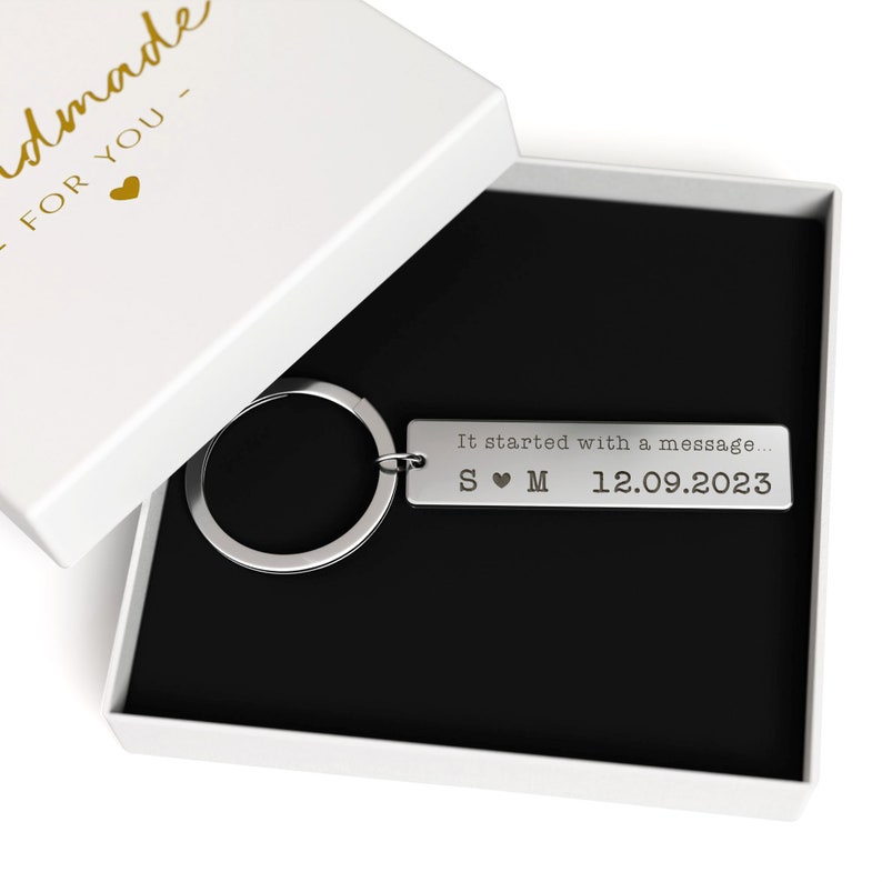 May include: A silver rectangular keychain with a keyring, presented in a white gift box with a black interior. The keychain is engraved with the text "It started with a message..." and initials S &hearts; M, along with the date 12.09.2023.