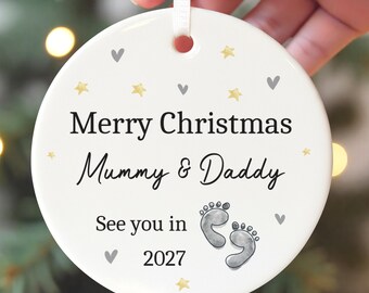Op de afbeelding: Een ronde witte keramische kerstornament met de zwarte tekst Merry Christmas Mummy & Daddy See you in 2027, versierd met kleine gouden sterretjes, grijze hartjes en een paar grijze babyvoetjes, opgehangen aan een wit lint.