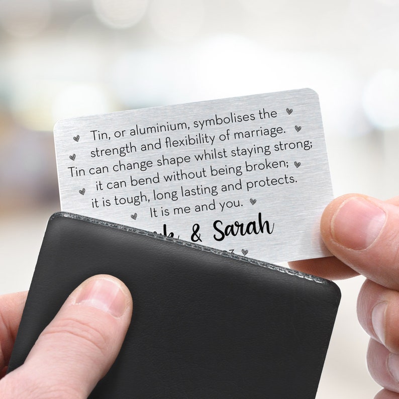 May include: A silver metal wallet card with engraved text. The card is held over a black wallet. The text reads: "Tin, or aluminium, symbolises the strength and flexibility of marriage... It is me and you." The names "Jake & Sarah" are also engraved.