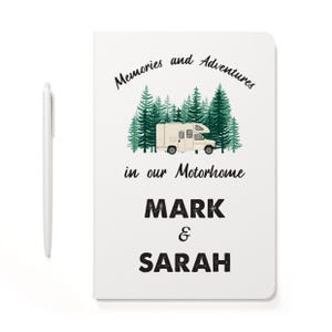 May include: A white notebook featuring a graphic of a beige motorhome parked in front of green pine trees. The text reads Memories and Adventures in our Motorhome, followed by the names MARK & SARAH. A white pen is positioned to the left.
