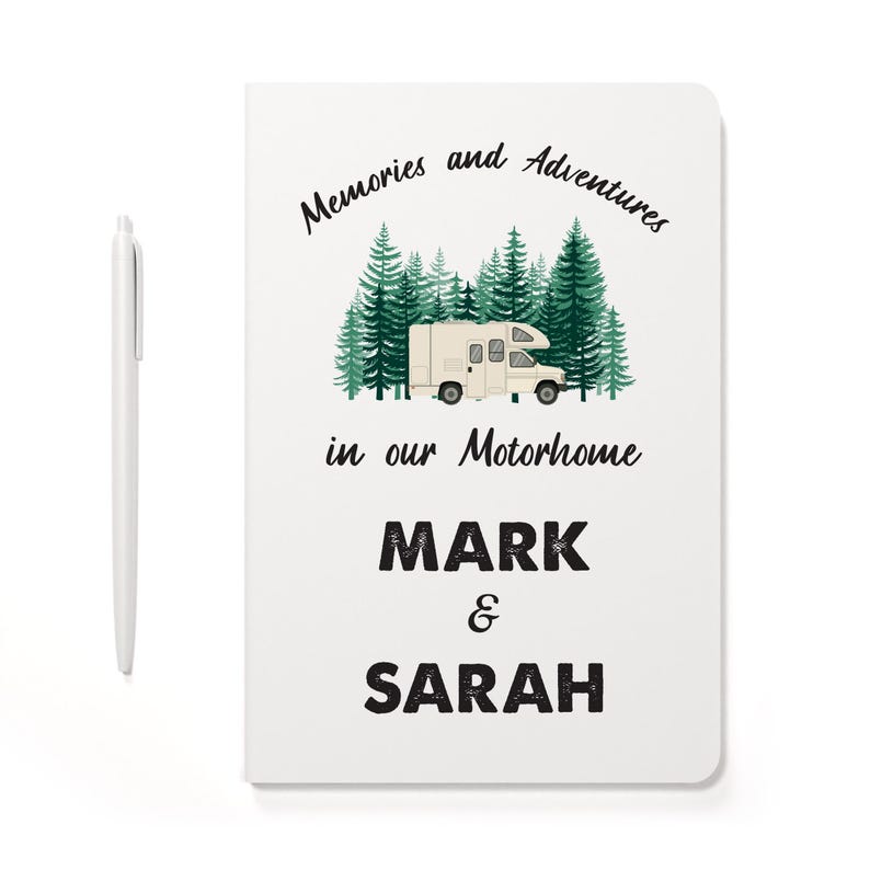 May include: A white notebook featuring a graphic of a beige motorhome parked in front of green pine trees. The text reads Memories and Adventures in our Motorhome, followed by the names MARK & SARAH. A white pen is positioned to the left.
