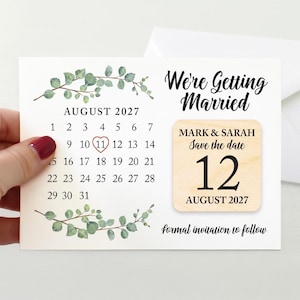 May include: A wedding save-the-date card with an August 2027 calendar, circled on the 11th. The card reads "We're Getting Married" and includes a wooden accent with the names "Mark & Sarah" and the date "12 August 2027". The design is decorated with green leaves.