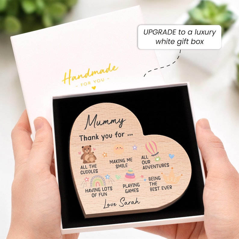 K&ouml;nnte beinhalten: Ein herzf&ouml;rmiges Holzschild mit der Aufschrift "Mummy, Thank you for..." und Illustrationen, pr&auml;sentiert in einer wei&szlig;en Geschenkbox. Das Schild enth&auml;lt S&auml;tze wie "All the cuddles" und "Love Sarah". Die Box ist mit "Handmade" beschriftet.