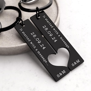 May include: Two black metal keychains with a heart shape cut out. The keychains are engraved with the text "It started with a message..." and the date "28.09.24". The keychains are also engraved with the initials "S&M".