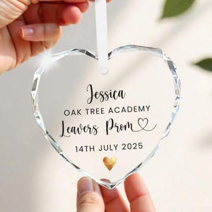 May include: Clear, heart-shaped ornament with a faceted design, held by hands. The ornament features the name "Jessica", "OAK TREE ACADEMY", "Leavers Prom", and the date "14TH JULY 2025", plus a gold heart.