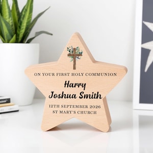 Op de afbeelding: Een lichtgekleurd houten aandenken in de vorm van een ster met de tekst "ON YOUR FIRST HOLY COMMUNION" en gepersonaliseerde details: "Harry Joshua Smith" en de datum "12 september 2026". Een kruis met bloemaccenten staat bovenaan.