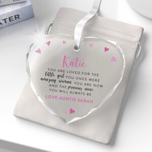 Puede incluir: Adorno de cristal transparente en forma de coraz&oacute;n, con una cinta blanca. El adorno presenta el nombre "Katie" en rosa, un mensaje cari&ntilde;oso y detalles de corazones rosas. Se presenta en una bolsa blanca con cord&oacute;n.