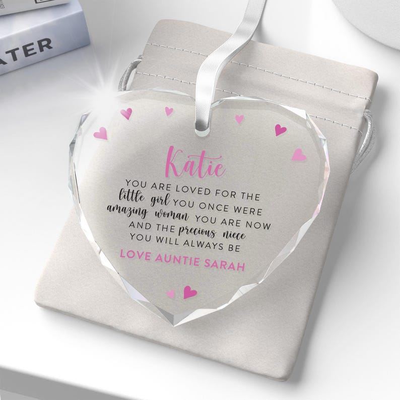Puede incluir: Adorno de cristal transparente en forma de coraz&oacute;n, con una cinta blanca. El adorno presenta el nombre "Katie" en rosa, un mensaje cari&ntilde;oso y detalles de corazones rosas. Se presenta en una bolsa blanca con cord&oacute;n.