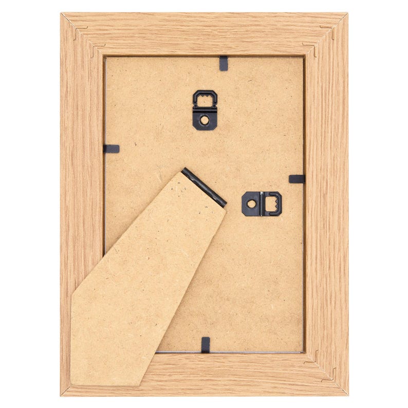 May include: Back of a rectangular wooden picture frame with a light brown finish. The frame has a cardboard backing, a hinged easel stand, and metal hanging hardware. The frame is designed to display photographs or artwork.