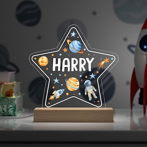 May include: A star-shaped night light with the name "HARRY" in white. The light features colorful illustrations of planets, stars, a rocket, and an astronaut. It is set on a wooden base and is designed for a child's room.