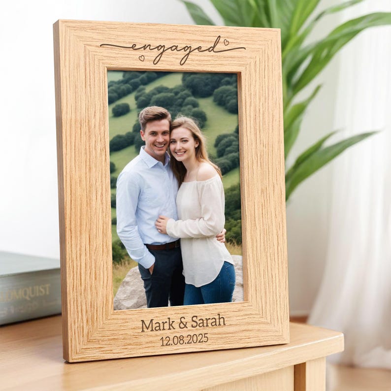 May include: A light-colored wooden picture frame with the word "engaged" in script above a photo of a smiling couple. The names "Mark & Sarah" and the date "12.08.2025" are engraved at the bottom of the frame.