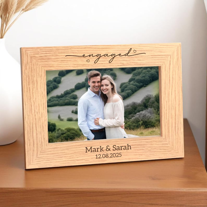 May include: A light-colored wooden picture frame with the word "engaged" in cursive script at the top. The frame holds a photo of a smiling couple. The names "Mark & Sarah" and the date "12.08.2025" are printed at the bottom.