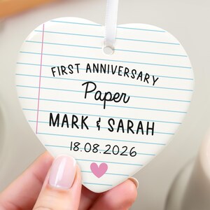 Puede incluir: Adorno de cer&aacute;mica en forma de coraz&oacute;n con dise&ntilde;o de papel rayado. El texto dice "FIRST ANNIVERSARY Paper MARK & SARAH 18.08.2026" con un coraz&oacute;n rosa. Se adjunta una cinta blanca para colgar.