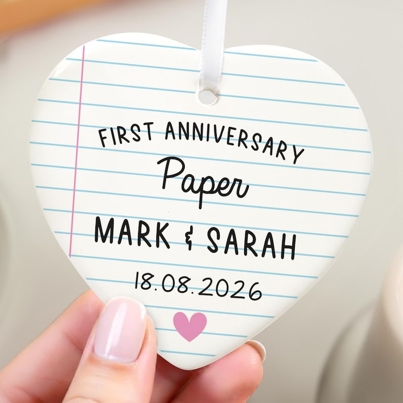 Puede incluir: Adorno de cer&aacute;mica en forma de coraz&oacute;n con dise&ntilde;o de papel rayado. El texto dice "FIRST ANNIVERSARY Paper MARK & SARAH 18.08.2026" con un coraz&oacute;n rosa. Se adjunta una cinta blanca para colgar.