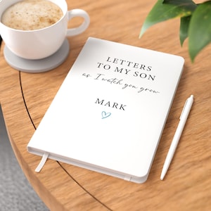 May include: A white journal with the black text "LETTERS TO MY SON as I watch you grow" and "MARK" printed on the cover, accompanied by a small blue heart icon. A white pen is placed next to the journal on a wooden surface near a coffee cup.