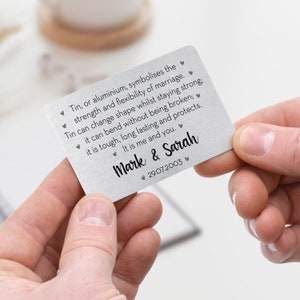 May include: A silver metal card held by two hands. The card has text that reads "Tin, or aluminium, symbolises the strength and flexibility of marriage." It is personalized with the names "Mark & Sarah" and the date "29.07.2003".