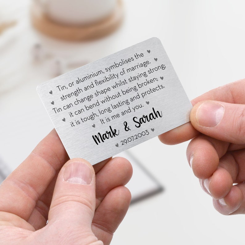 May include: A silver metal card held by two hands. The card has text that reads "Tin, or aluminium, symbolises the strength and flexibility of marriage." It is personalized with the names "Mark & Sarah" and the date "29.07.2003".