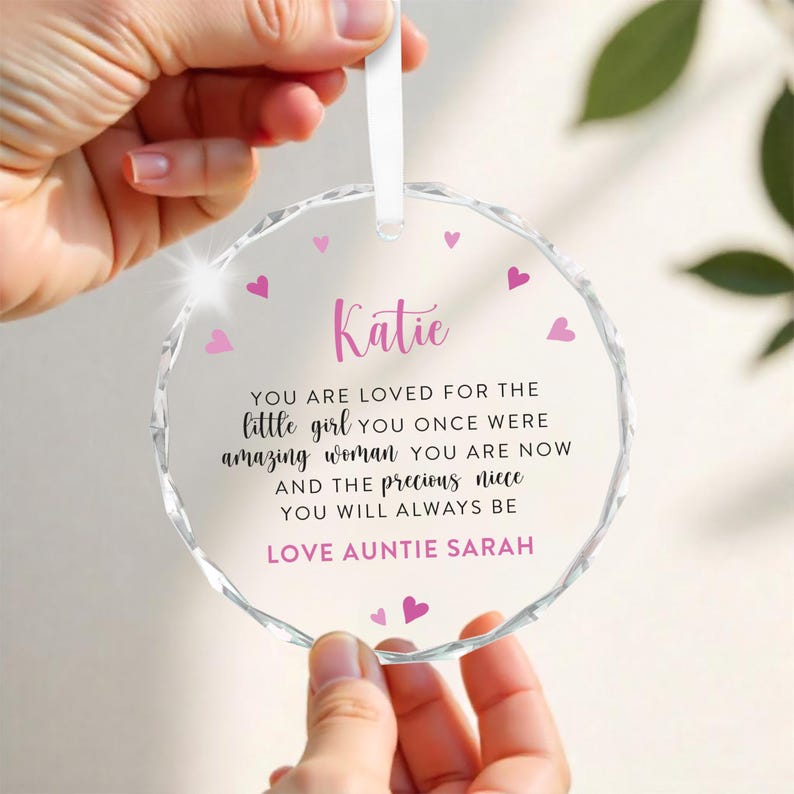 Puede incluir: Un adorno de cristal transparente y redondo con una cinta blanca. El adorno tiene un borde facetado y corazones rosas. El nombre "Katie" est&aacute; escrito en rosa, junto con un mensaje cari&ntilde;oso de "T&iacute;a Sarah".