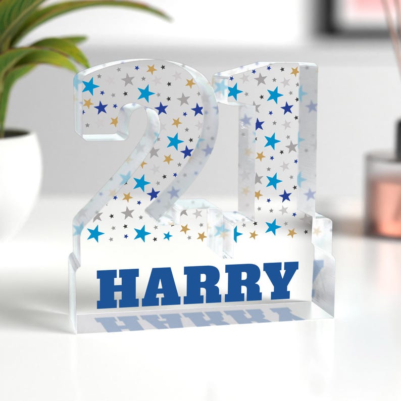 Puede incluir: Un n&uacute;mero 21 de acr&iacute;lico transparente con un patr&oacute;n de estrellas azules, doradas y grises. El nombre "HARRY" est&aacute; impreso en letras azules en la base. Un art&iacute;culo decorativo de cumplea&ntilde;os.