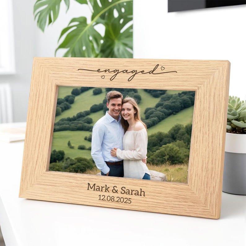 May include: A light-colored wooden picture frame with the word "engaged" in cursive script at the top. The frame holds a photo of a couple and is inscribed with "Mark & Sarah 12.08.2025" at the bottom.