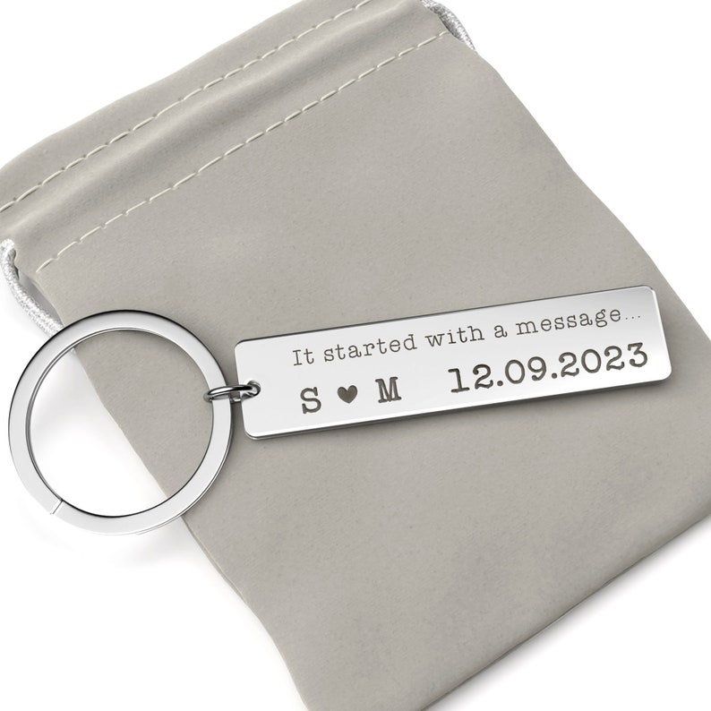 May include: A silver rectangular keychain with a key ring, resting on a gray pouch. The keychain has the engraved text "It started with a message..." and the date "12.09.2023". The initials "S &hearts; M" are also engraved.