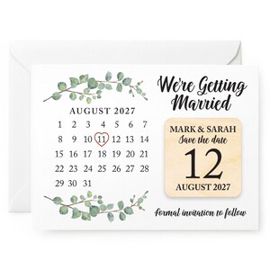 May include: A white save-the-date card featuring a calendar for August 2027, with the 11th circled in red. The card reads "We're Getting Married" and has a wooden accent with the date August 12, 2027, for Mark & Sarah. Includes eucalyptus.
