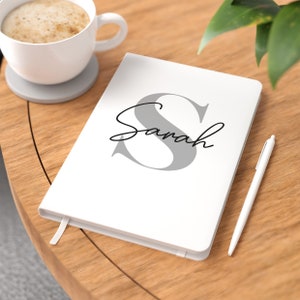 May include: White notebook with a large grey letter "S" and the name "Sarah" written in black cursive. A white pen is lying on a wooden table next to the notebook.