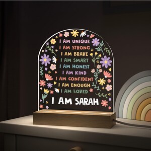 May include: A decorative night light with a wooden base and a clear acrylic panel. The panel features colorful text that reads "I AM UNIQUE, I AM STRONG, I AM BRAVE, I AM SMART, I AM HONEST, I AM KIND, I AM CONFIDENT, I AM ENOUGH, I AM LOVED, I AM SARAH" with floral accents.