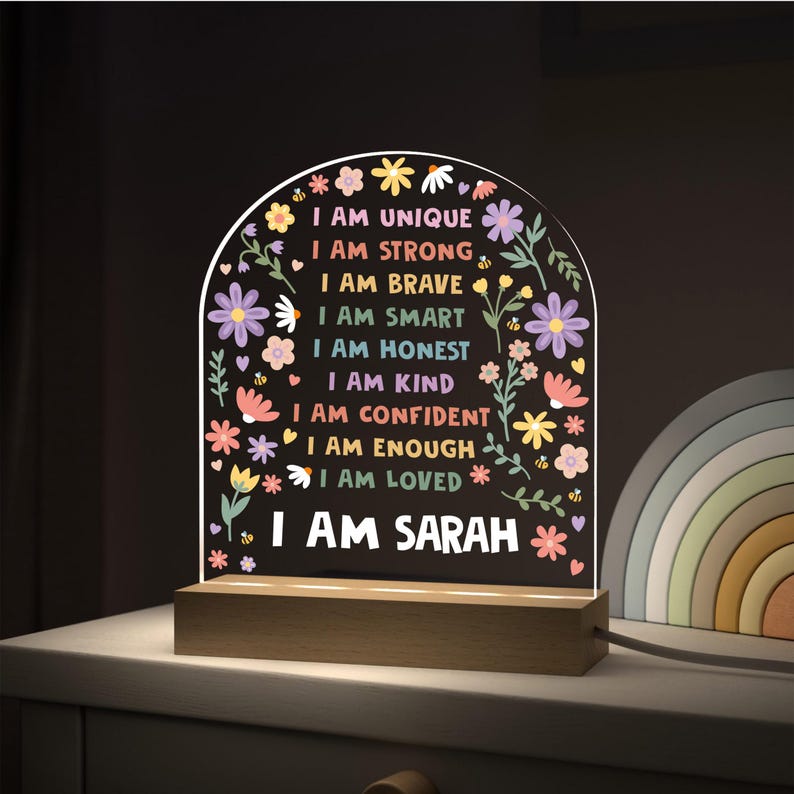 May include: A decorative night light with a wooden base and a clear acrylic panel. The panel features colorful text that reads "I AM UNIQUE, I AM STRONG, I AM BRAVE, I AM SMART, I AM HONEST, I AM KIND, I AM CONFIDENT, I AM ENOUGH, I AM LOVED, I AM SARAH" with floral accents.