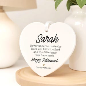 Op de afbeelding: Een witte, hartvormige keramische plaquette met de naam "Sarah" in elegant schrift. De plaquette bevat de tekst "Never underestimate the lives you have touched...Happy Retirement". Een wit lint is bevestigd om op te hangen.