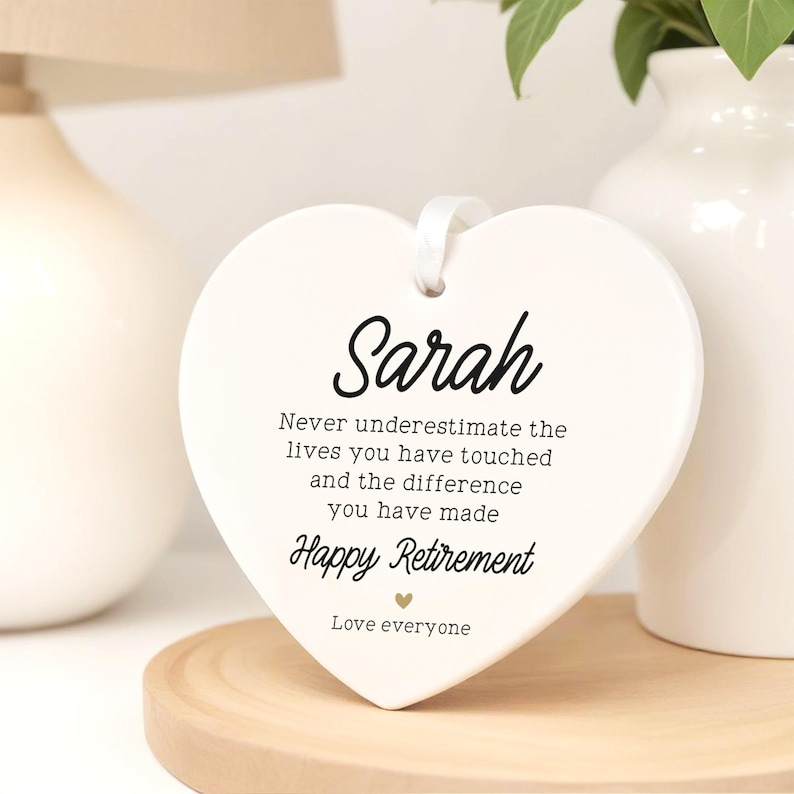 Op de afbeelding: Een witte, hartvormige keramische plaquette met de naam "Sarah" in elegant schrift. De plaquette bevat de tekst "Never underestimate the lives you have touched...Happy Retirement". Een wit lint is bevestigd om op te hangen.