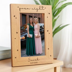 May include: A light-coloured wooden picture frame with a vertical orientation. The frame is engraved with the text "prom night" at the top, "Sarah" at the bottom, and the date "12.07.2026". Small heart and star icons are etched along the sides.