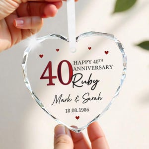May include: A heart-shaped, faceted glass ornament with a white ribbon. The ornament features the text "40 Ruby Anniversary" in red and black, along with the names "Mark & Sarah" and the date "18.08.1986". Small red hearts are scattered around the text.