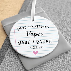 Puede incluir: Adorno redondo de cer&aacute;mica blanca con dise&ntilde;o de papel rayado. El texto dice "FIRST ANNIVERSARY PAPER MARK & SARAH 18.08.26" con un coraz&oacute;n rosa. Una cinta blanca est&aacute; adjunta para colgar. El adorno est&aacute; sobre una bolsa gris.