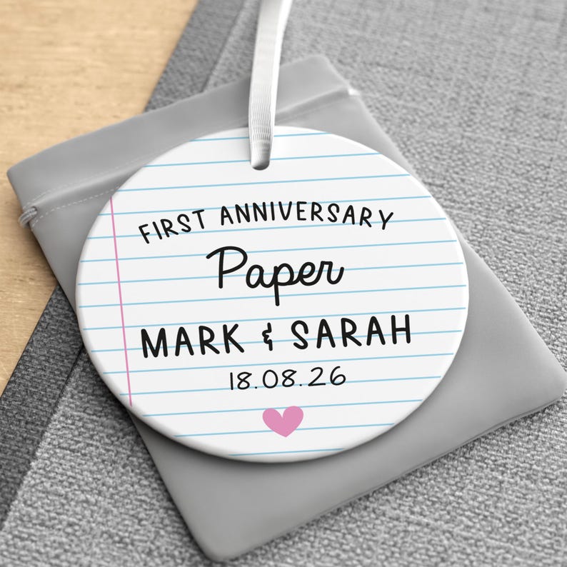 Puede incluir: Adorno redondo de cer&aacute;mica blanca con dise&ntilde;o de papel rayado. El texto dice "FIRST ANNIVERSARY PAPER MARK & SARAH 18.08.26" con un coraz&oacute;n rosa. Una cinta blanca est&aacute; adjunta para colgar. El adorno est&aacute; sobre una bolsa gris.