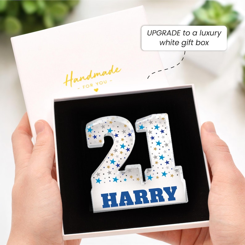 Puede incluir: Una caja de regalo blanca con el texto dorado "Handmade - FOR YOU" contiene el n&uacute;mero "21" de acr&iacute;lico transparente decorado con estrellas azules y doradas. El nombre "HARRY" est&aacute; impreso en azul. La caja est&aacute; abierta, revelando un interior negro.