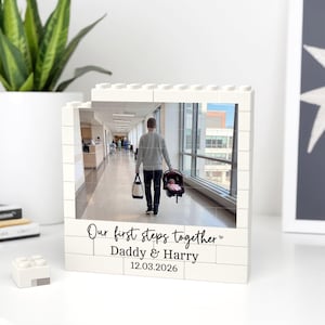 May include: A white brick-style photo frame displaying a photograph of a person walking down a corridor with a baby in a car seat. The frame is inscribed with "Our first steps together Daddy & Harry 12.03.2026".