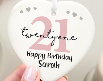 May include: A heart-shaped, white ceramic ornament with a pink "21" and the words "twentyone" and "Happy Birthday Sarah" in black script. Small gray hearts are scattered across the top. A white ribbon is attached for hanging.