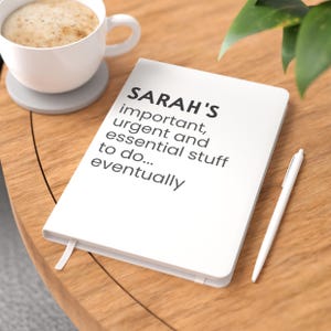 May include: A white hardback notebook featuring the black printed text: SARAH'S important, urgent and essential stuff to do... eventually. The notebook includes a white ribbon bookmark and is accompanied by a slim white pen.