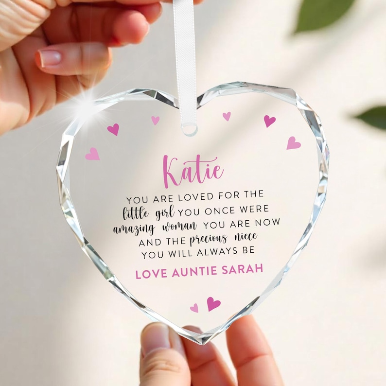 Puede incluir: Un adorno transparente en forma de coraz&oacute;n con un dise&ntilde;o facetado. El adorno tiene una cinta blanca para colgar y presenta el nombre "Katie" en escritura rosa, junto con un mensaje de amor y peque&ntilde;os corazones rosas.