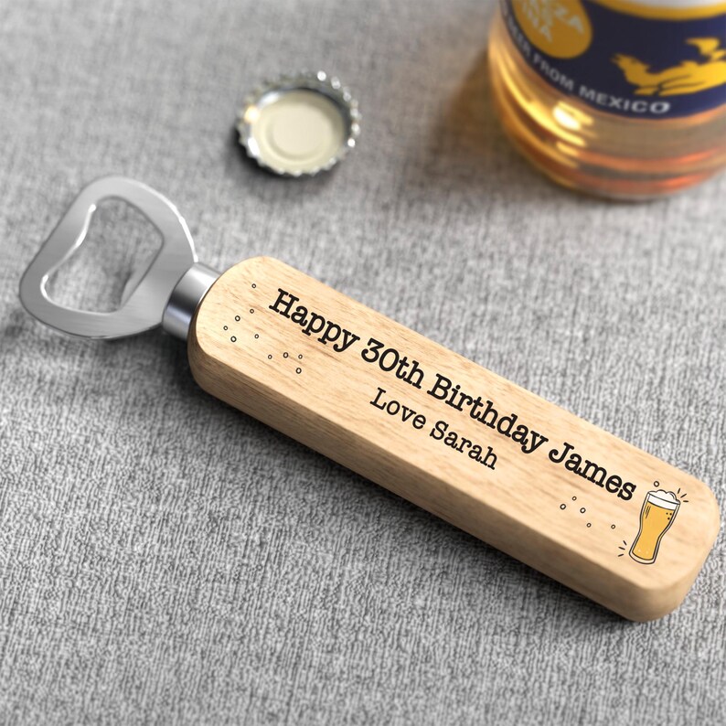 Puede incluir: Un abridor de botellas de madera con cabezal de acero inoxidable. El abridor est&aacute; grabado con "Happy 30th Birthday James Love Sarah" y un gr&aacute;fico de vaso de cerveza. Una chapa de botella y una botella de cerveza est&aacute;n en el fondo.