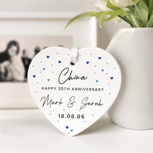 Op de afbeelding: Een wit hartvormig keramisch ornament met blauwe hart- en stipaccenten. De tekst luidt China, HAPPY 20TH ANNIVERSARY, Mark & Sarah en 18.08.06. Een wit lint is door een gat aan de bovenkant geregen om het ornament op te hangen.
