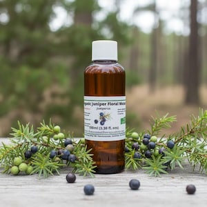 Puede incluir: Una botella de vidrio marrón con tapa blanca, etiquetada como "Organic Juniper Floral Water Juniperus 100ml (3.38 fl. oz.)", se encuentra entre ramas de enebro con bayas verdes y azules. El fondo es un bosque borroso.