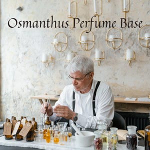 May include: An individual in a white shirt and suspenders is working with various glass bottles and beakers. The text "Osmanthus Perfume Base" is displayed above. The scene suggests a laboratory or workshop setting, with a focus on fragrance creation.
