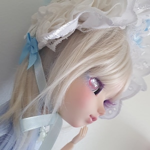 Può includere: Una bambola con lunghi capelli bianchi e occhi azzurri. Indossa un cappello di pizzo bianco e un vestito azzurro chiaro con un'applicazione di pizzo bianco.